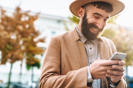 Attractive smiling young man wearing autumn coat and hat walking outdoors at the city street, holding mobile phoneの写真素材