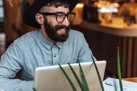 Photo of joyful young man wearing eyeglasses using laptop and smiling while sitting at table in cafe indoorsの写真素材