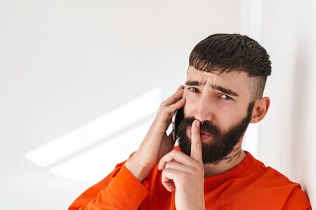 Image of young bearded man with nose jewelry wearing orange shirt talking on smartphone and holding finger on lips over white wall indoorsの写真素材