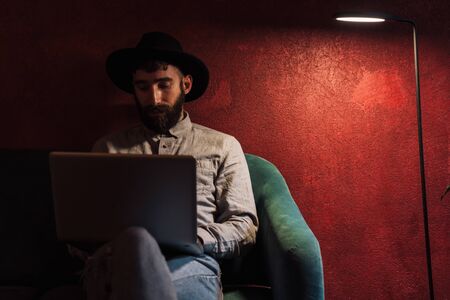 Photo of focused young man wearing hat using laptop while sitting in armchair at cafe indoorsの写真素材