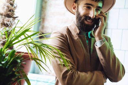 Image of joyful stylish young man wearing hat smiling and using cellphone in cafeの写真素材