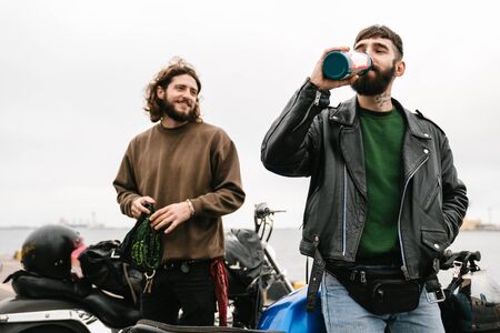 Photo of bearded young men bikers drinking from bottle and smiling while standing with their bikes...