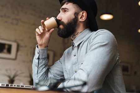 Photo of thinking young man wearing hat using laptop and drinking coffee while sitting at table in...
