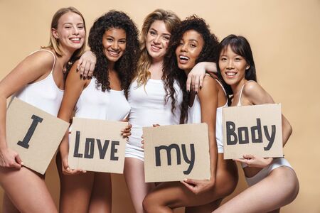 Image of cheerful multinational women in underclothes hugging and holding placards isolated over beigeの写真素材