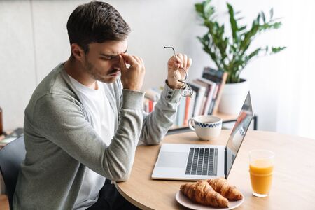 Image of tired young man with eyeglasses expressing fatigue while working on laptop at homeの写真素材