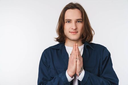 A pleased caucasian man with long hair posing and holding palms together isolated over white