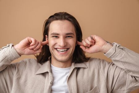 A joyful young man with red hair smiling and plugging his ears isolated over beigeの写真素材