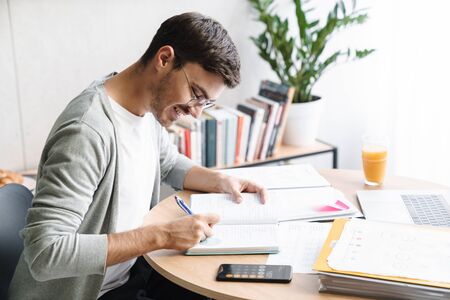 Image of happy young man in eyeglasses doing calculations while working on laptop at home