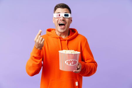 A surprised emotional young man posing wearing 3d glasses eat popcorn.の写真素材