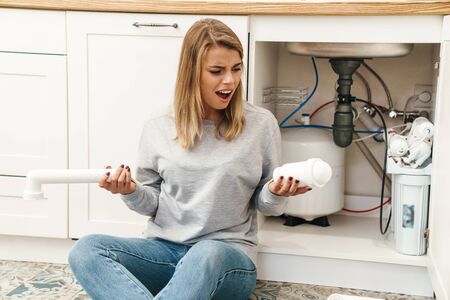 Photo of displeased young blonde woman with plumbing pipes while sitting on floor near kitchen sinkの写真素材