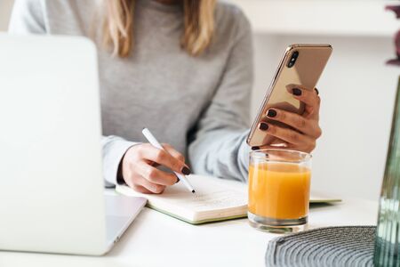 Cropped photo of young woman in eyeglasses making notes in planner while using cellphone at living roomの写真素材