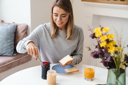 Photo of cheerful young woman in eyeglasses eating toasts while having breakfast at living roomの写真素材