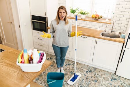 Photo of irritated woman housewife in gloves screaming while mopping floor at modern kitchen