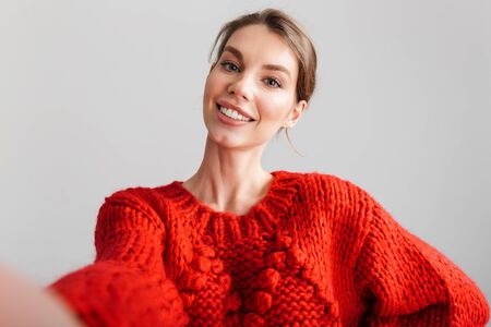 Image of young cheerful woman in red sweater smiling and taking selfie photo isolated over white backgroundの写真素材