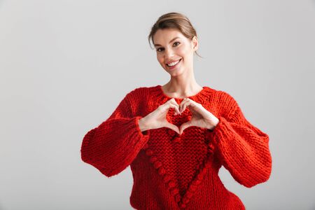 Image of young cheerful woman in red sweater making heart gesture with fingers isolated over white backgroundの写真素材