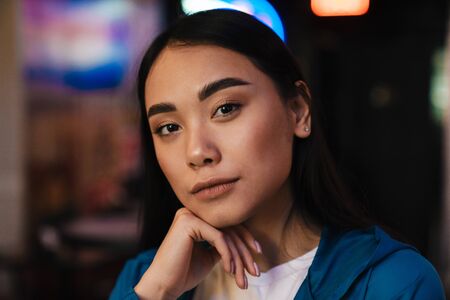 Photo of serious young asian woman with brunette hair looking at camera while posing in cafeの写真素材