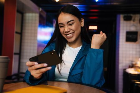 Photo of excited asian woman playing video game on mobile phone and making winner gesture while sitting in cafeの写真素材
