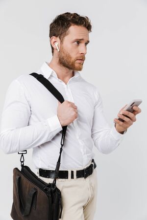 Image of thinking blonde man using cellphone and wireless earphones isolated over white backgroundの写真素材