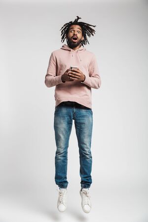 Full length portrait of a young african man wearing hoodie jumping isolated over white background, holding mobile phoneの写真素材