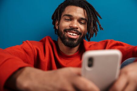 Close up portrait of a smiling young african man wearing pullover sitting isolated over blue background, using mobile phoneの写真素材