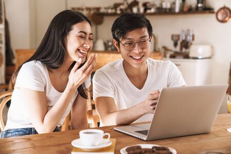 Image of multicultural happy couple smiling and using laptop while sitting at table in cozy kitchenの写真素材