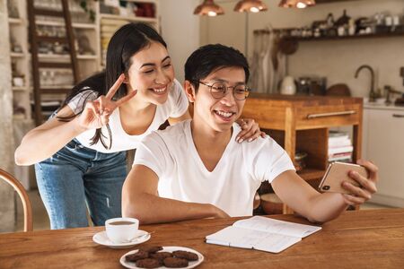 Image of multicultural smiling couple taking selfie photo on smartphone and gesturing peace sign in cozy kitchenの写真素材