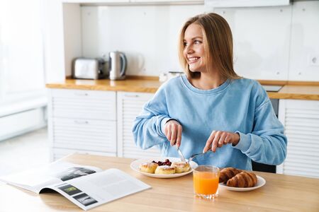 Image of cheerful young woman with blonde hair reading magazine while having breakfast at modern kitchenの写真素材