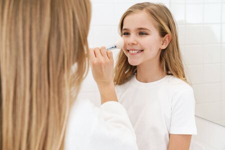 Photo of pleased caucasian mother and daughter smiling while doing makeup in white bathroomの写真素材