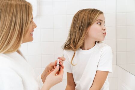 Photo of joyful caucasian mother and daughter smiling while doing makeup in white bathroomの写真素材