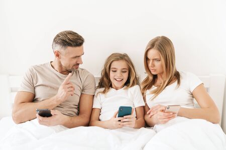 Photo of young caucasian family expressing shock and using cellphones while sitting in white bad...