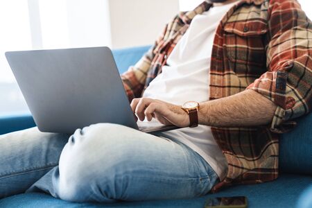 Portrait of young caucasian man wearing plaid shirt using laptop while sitting on sofa at homeの写真素材