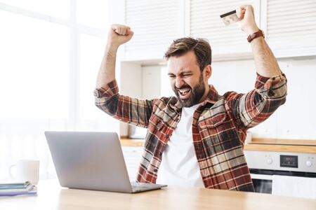 Portrait of excited bearded man holding credit card and making winner gesture while working with laptop at kitchenの写真素材