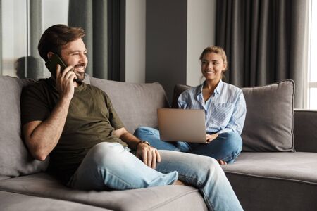 Photo of young caucasian joyful couple using laptop and mobile phone while sitting at living roomの写真素材