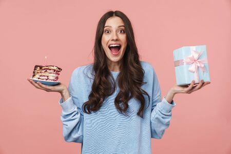 Image of delighted young woman with long brown hair holding birthday cake and present box isolated...