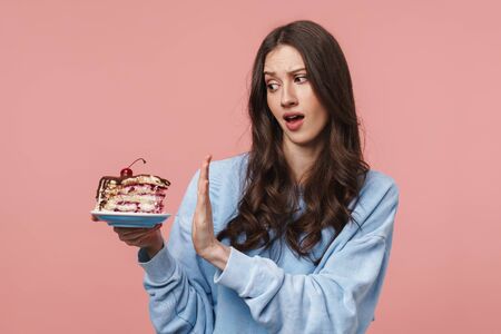 Image of displeased brunette woman doing refusal gesture while holding piece of cake isolated over pink backgroundの写真素材
