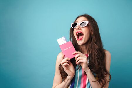Photo of astonished young woman in sunglasses posing with passport and tickets isolated over blue wallの写真素材