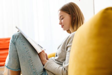 Beautiful smiling young blonde woman wearing casual clothes resting on a couch at home, reading a magazineの写真素材
