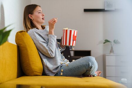 Side view of a beautiful shocked young blonde woman wearing casual clothes resting on a couch at home, watching movie, eating popcornの写真素材