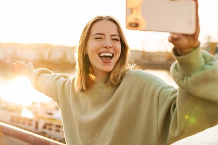 Portrait of cheerful woman smiling and taking selfie on cellphone while walking on promenadeの写真素材