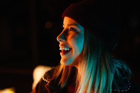 Photo of beautiful joyful woman smiling and looking aside while walking at park with neon lightingの写真素材