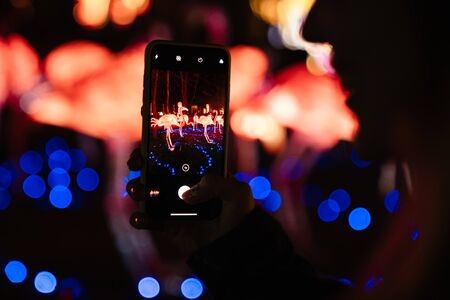 Photo of young woman taking photo on smartphone while walking at park with neon lightingの写真素材