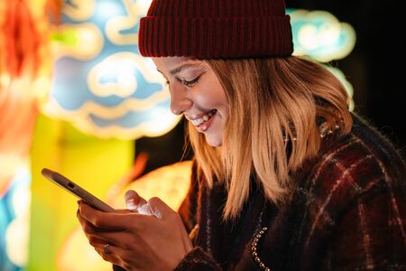 Photo of cheerful nice woman smiling and typing on mobile phone while walking at park with neon lightingの写真素材