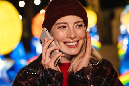 Photo of joyful nice woman smiling and talking on mobile phone while walking at park with neon lightingの写真素材