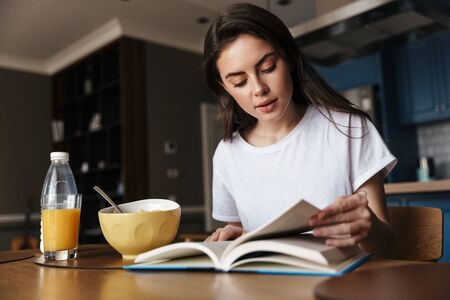 Attractive young brunette woman having a healthy breakfast reading a book while sitting at the kitchen tableの写真素材