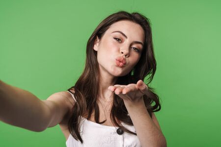 Image of young attractive woman blowing air kiss and taking selfie photo isolated over green backgroundの写真素材