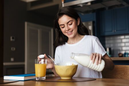 Attractive smiling young brunette woman having a healthy breakfast while sitting at the kitchen table, pouring milk in a cerealの写真素材
