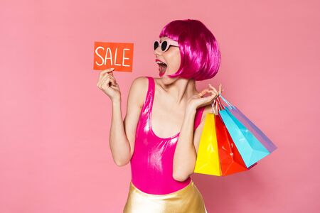 Image of young happy girl wearing wig holding colorful shopping bags and sale card isolated over pink backgroundの写真素材