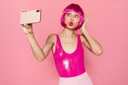 Image of joyful woman in headphone making kiss lips and taking selfie on cellphone isolated over pink backgroundの写真素材