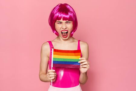 Image of displeased woman in wig screaming and holding rainbow flag isolated over pink backgroundの写真素材