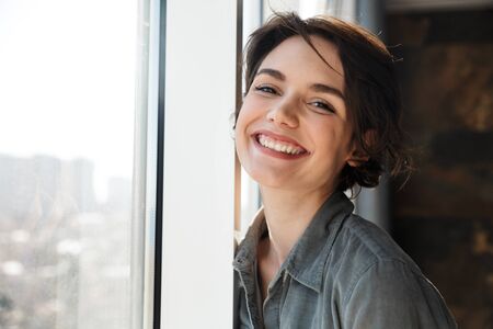 Image of beautiful young joyful woman smiling and looking at camera while standing near window
